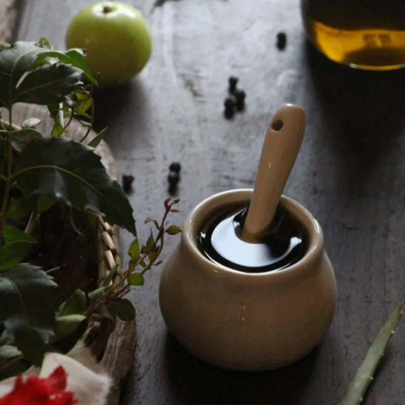 Small ceramic container with hair oil on a wooden surface with ayurvedic herbs.